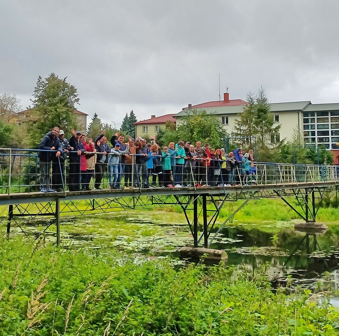 Grupa uczestników zajęć wraz z opiekunami stojąca na kładce dla pieszych nad rzeczką Kałamanka. Na lustrze wody widoczna roślinność wodna. W tle widoczny budynek WTZ.