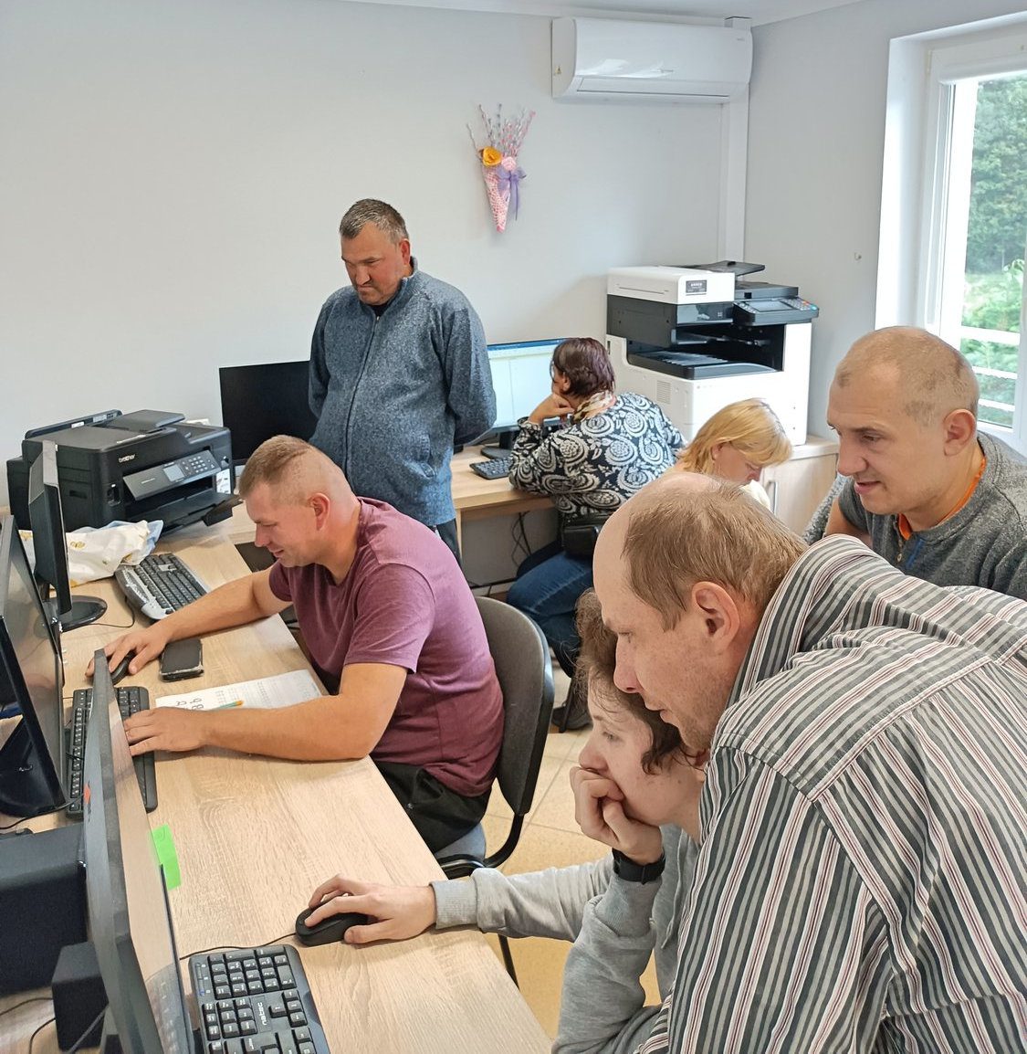 Uczestnicy zajęć pracują przy stanowiskach komputerowych. Część osób pracuje w grupach a część pojedyńczo. W tle widoczne dwa urządzenia wielofunkcyjne.